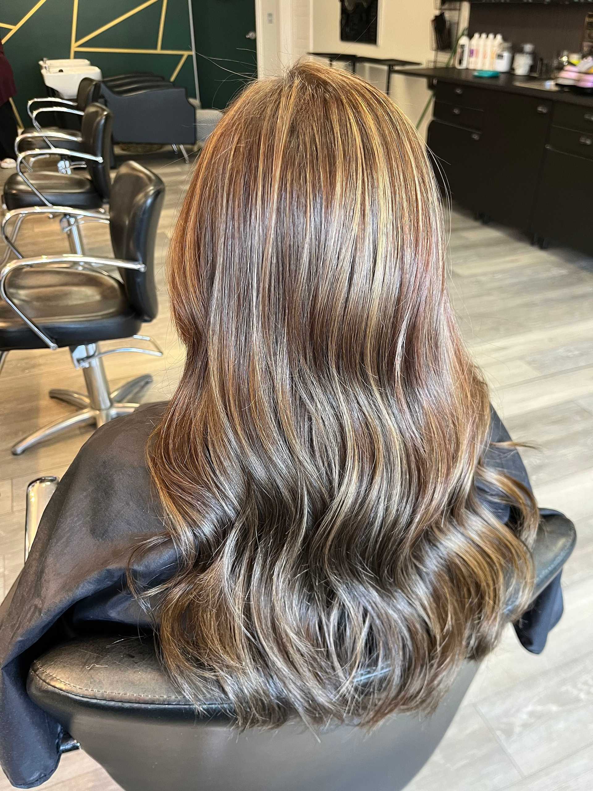 Woman with wavy, highlighted hair in a salon chair, modern decor in the background.